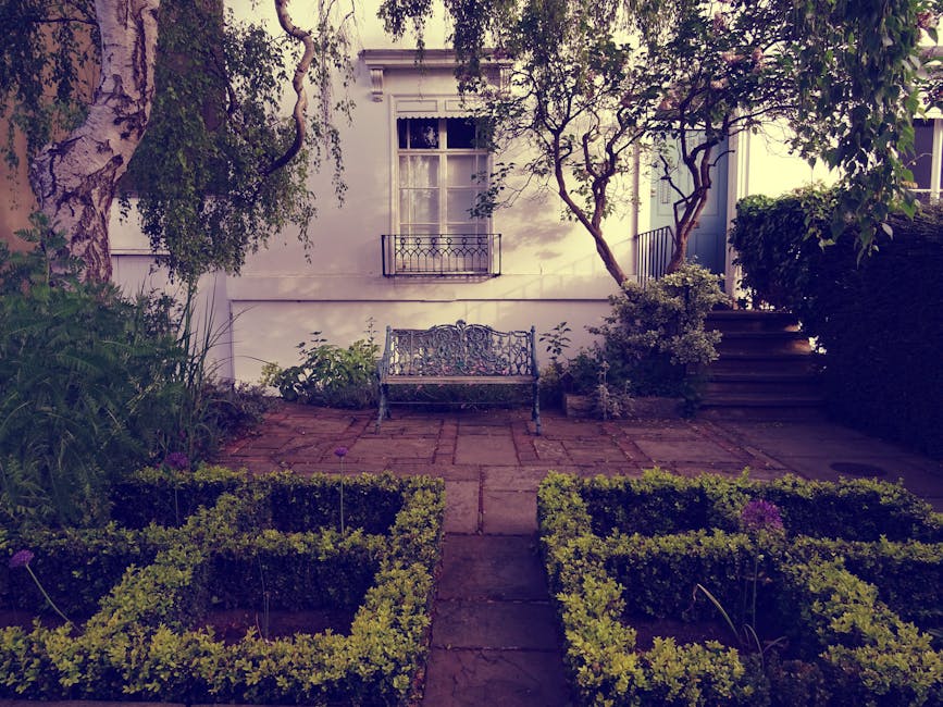 A small, well-maintained garden courtyard featuring a paved stone patio with a decorative wrought iron bench positioned against the white exterior wall of a building. The garden is bordered by neatly
