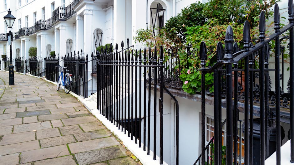 A small, well-maintained garden courtyard featuring a paved stone patio with a decorative wrought iron bench positioned against the white exterior wall of a building. The garden is bordered by neatly